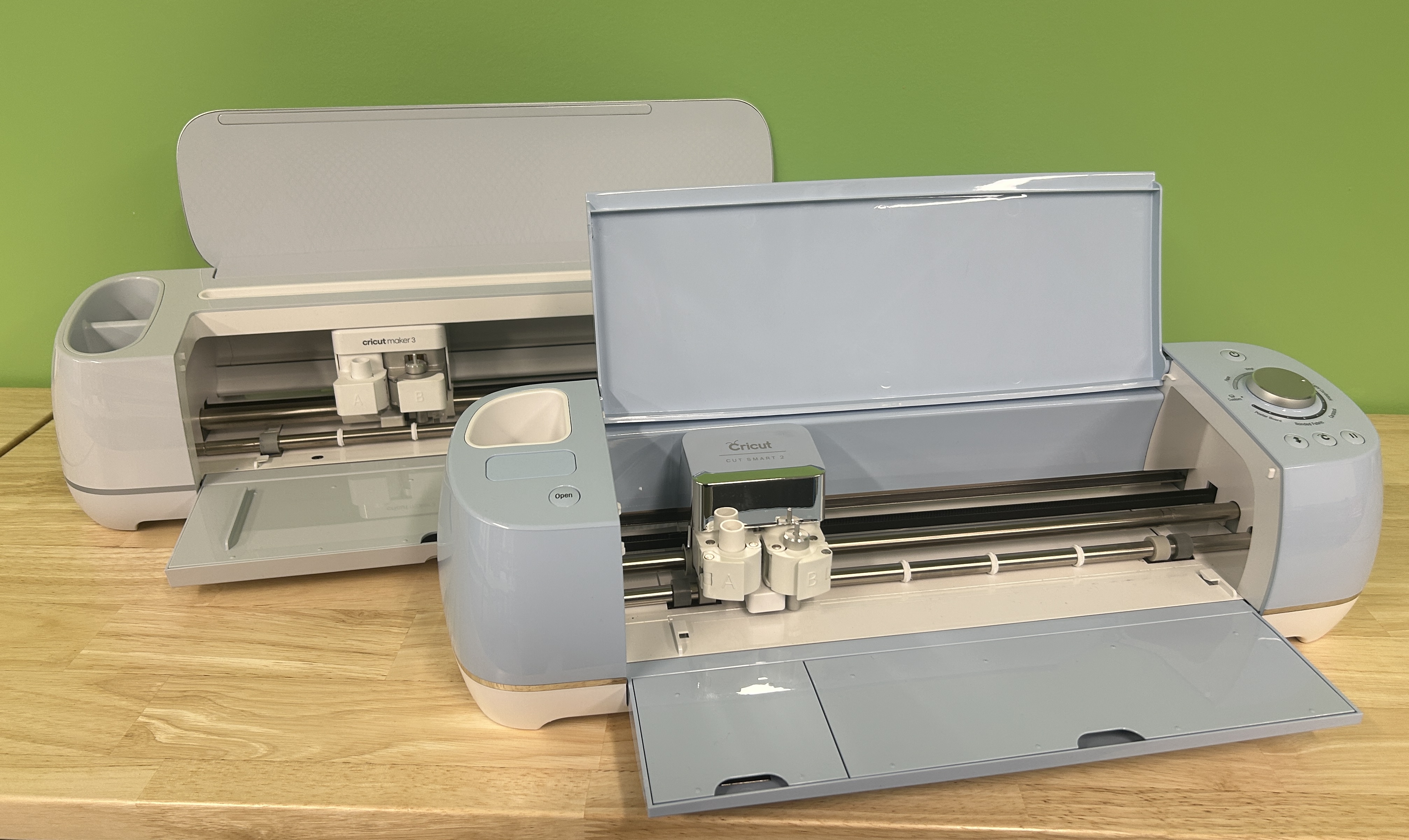 two Cricut machines, one white Maker 3 and one blue Explore Air 2, sitting on a table against a green wall