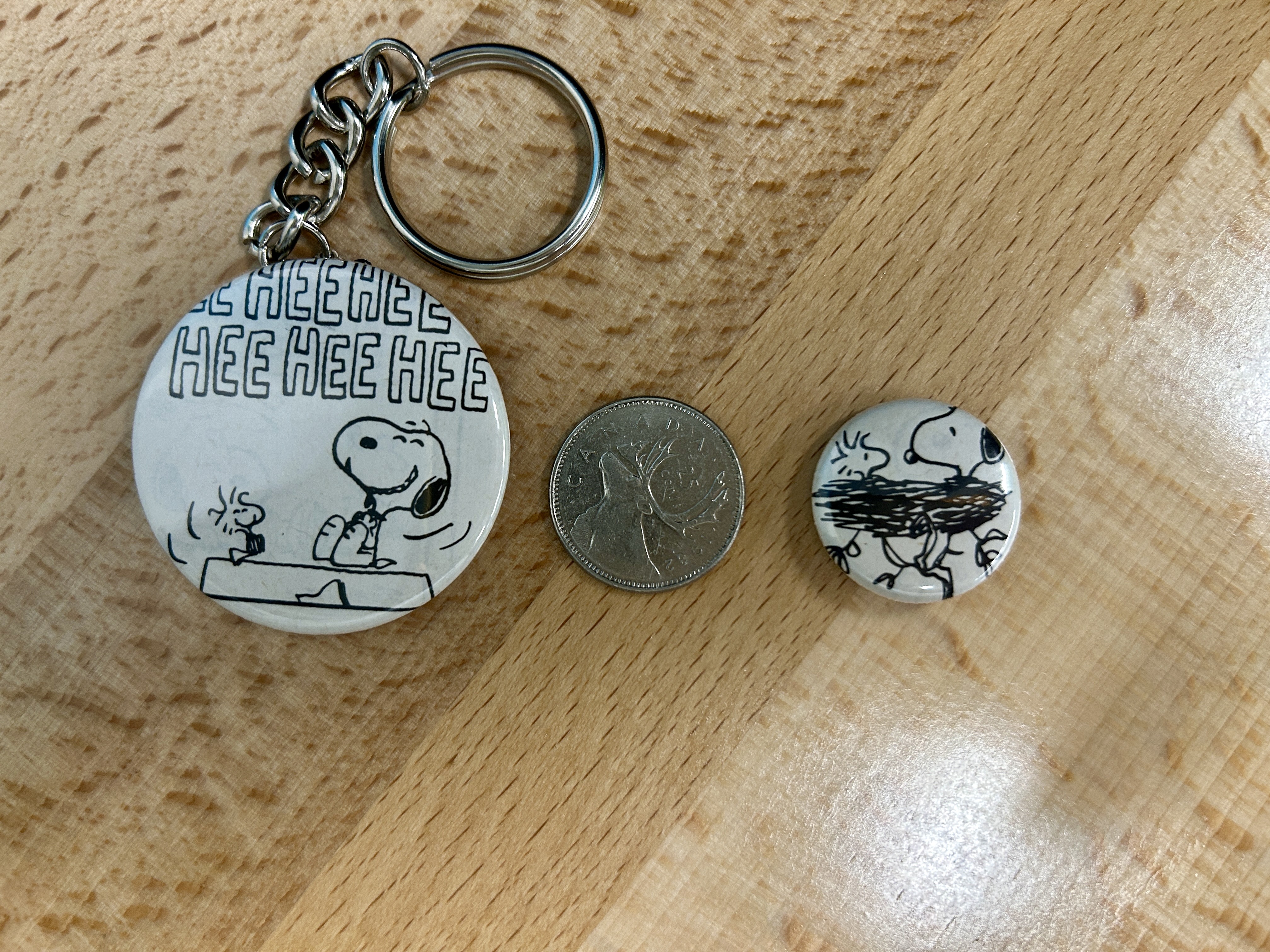 a 1.75 inch pin with a laughing Snoopy and Woodstock on it. To the right is a Canadian quarter. To the right of the quarter is a 1 inch pin with Snoopy and Woodstock sitting in a nest on it