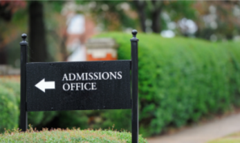 Green hedge next to concrete sidewalk in background with a black sign with white text "Admissions Office" and arrow pointing left