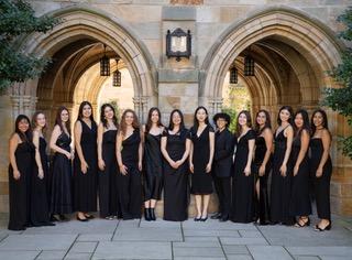 15 women dressed in black formal attire standing in from of two arches at Yale.