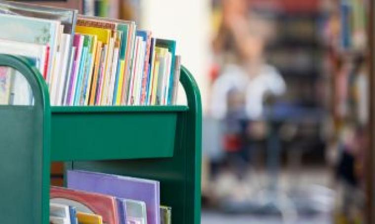 Green book cart holding a full shelf of picture books on the left half of the image.  Right half of the image is a blurred library background